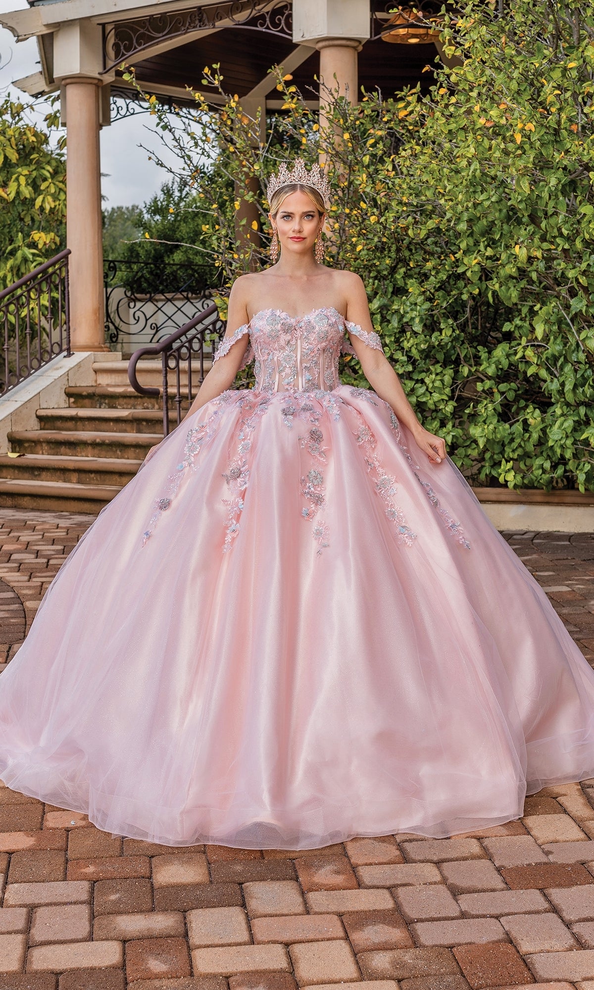 A person wearing the Quinceanera Dress 1836 By Dancing Queen, a blush pink ball gown with floral embellishments and a crown, stands outside on a paved area near greenery and a staircase.